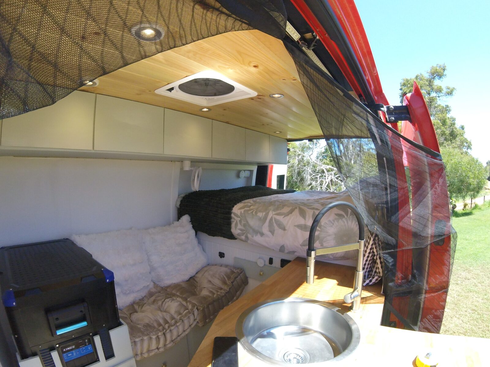 Inside the finished van looking out through the open side door and its fly screen, showing the bed, seating and sink